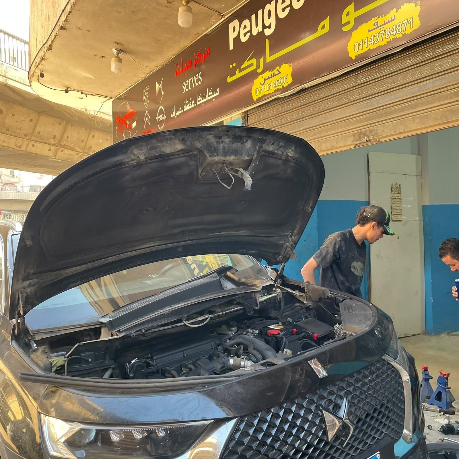 Two mechanics working on a black DS with its bonnet open outside the workshop.