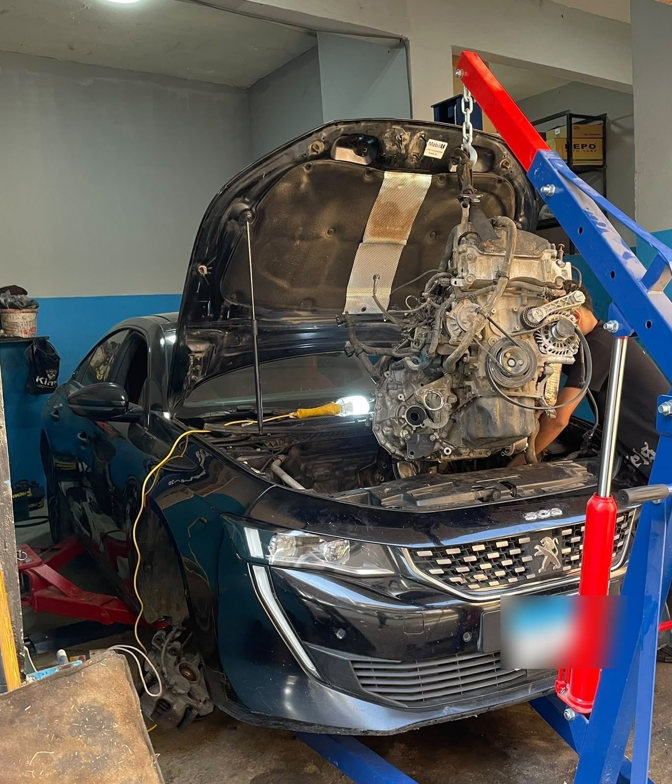 An engine hanging from a red and blue engine crane above the front of a black Peugeot 508 inside the workshop.