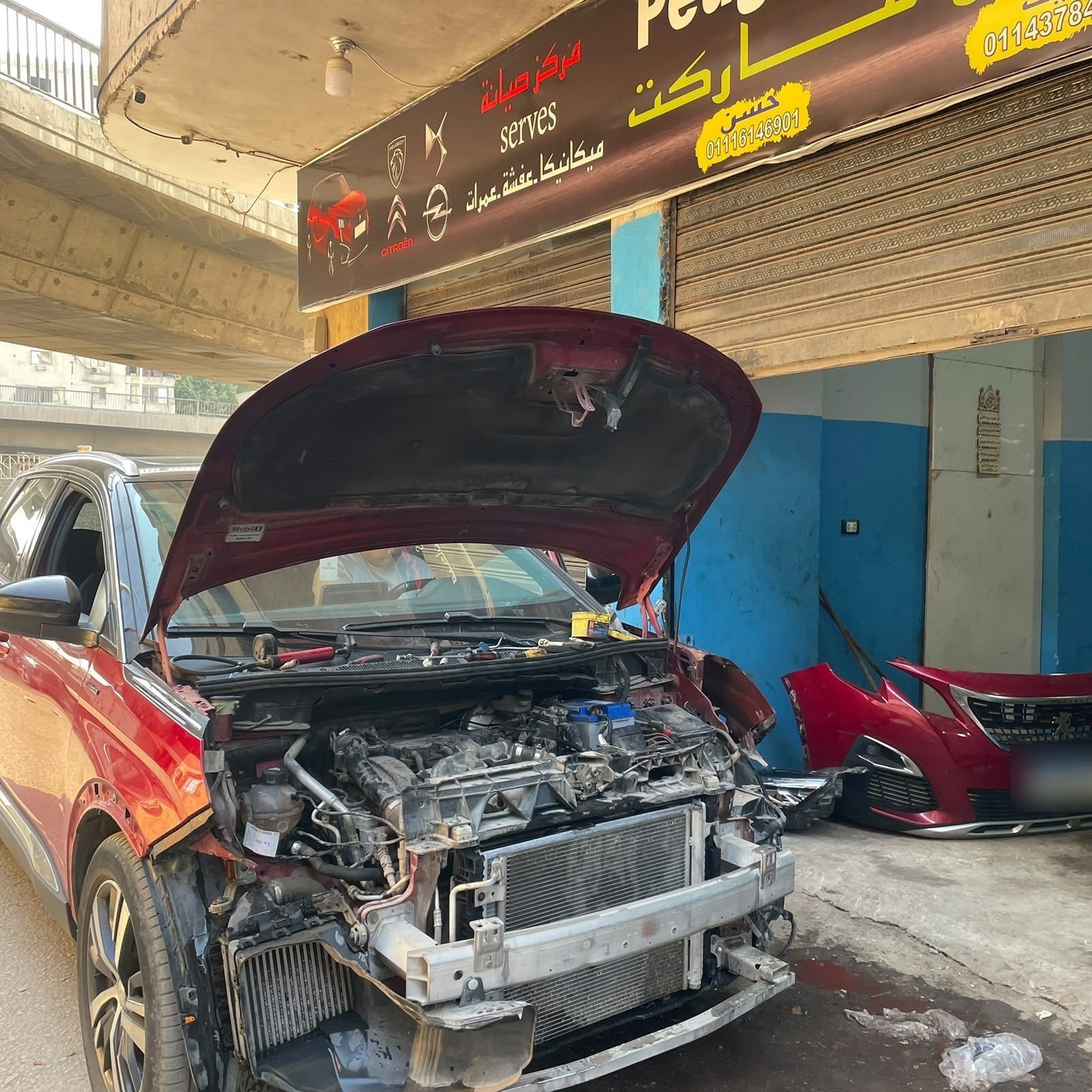 A red Peugeot outside the workshop with its front end removed and the front bumper on the ground beside it.