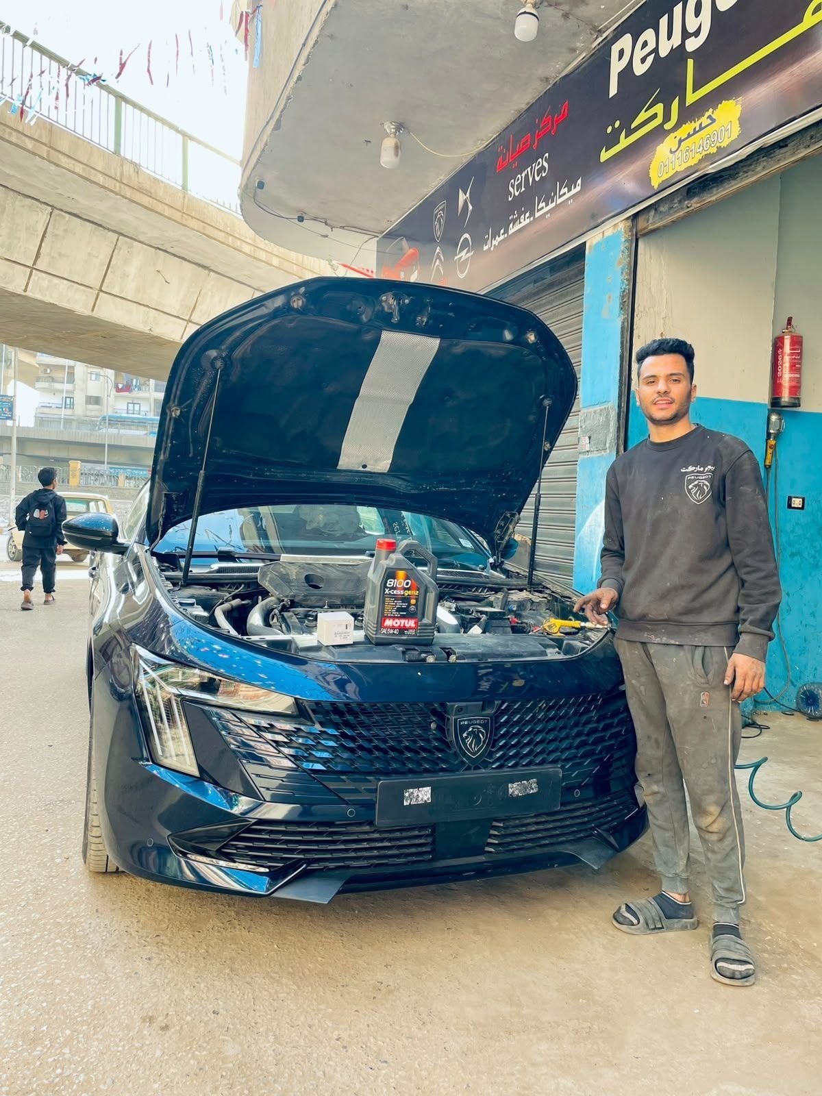 A mechanic beside a blue Peugeot with its bonnet open, with an oil bottle and a filter box on the engine.