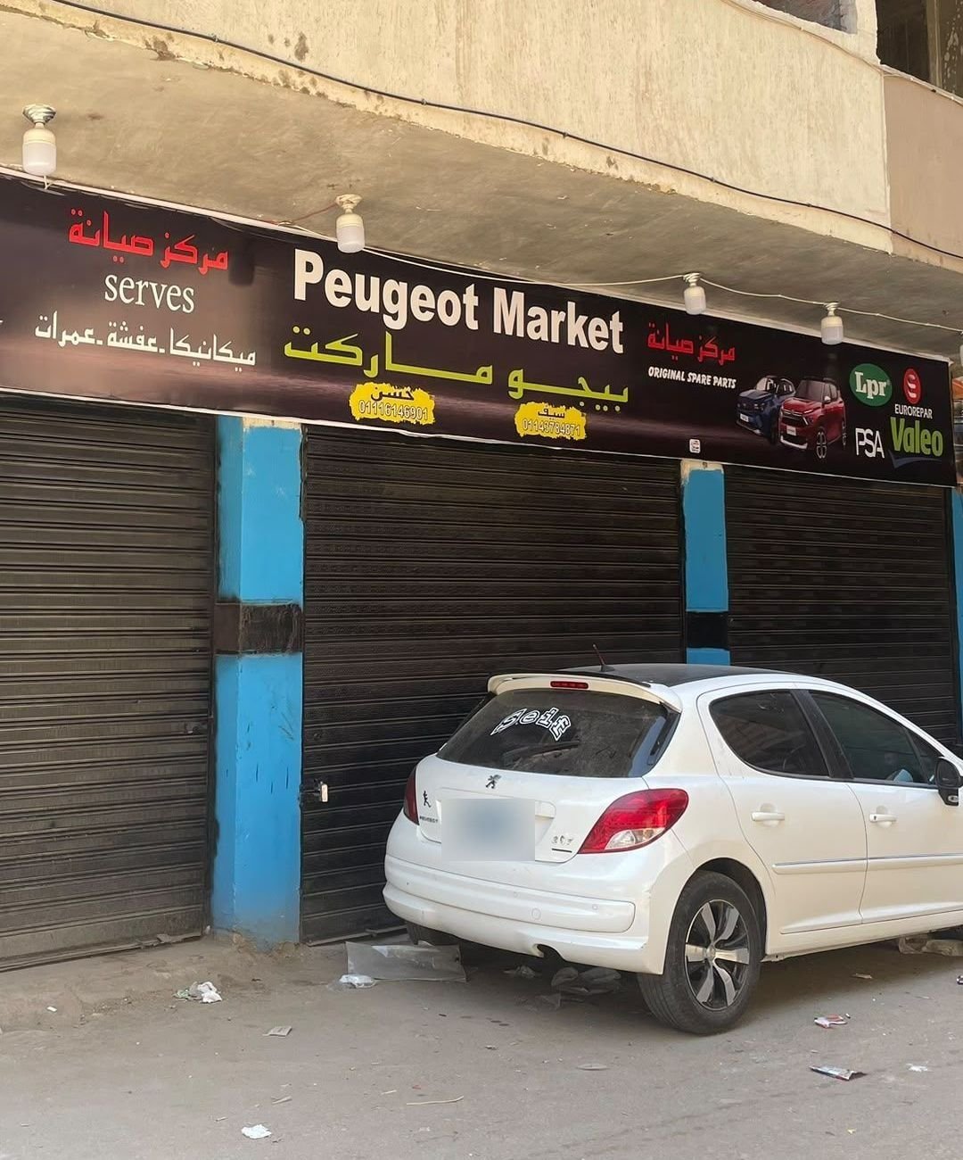 The Peugeot Market shopfront with its shutters down: a long black sign above blue pillars, with a white Peugeot 207 parked in front.