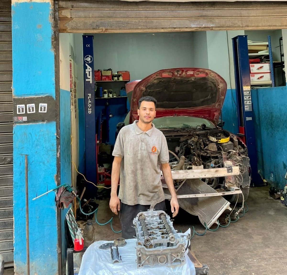 A mechanic at the workshop entrance with a cylinder head on the table in front of him and a red car with its bonnet open between the lift posts behind.